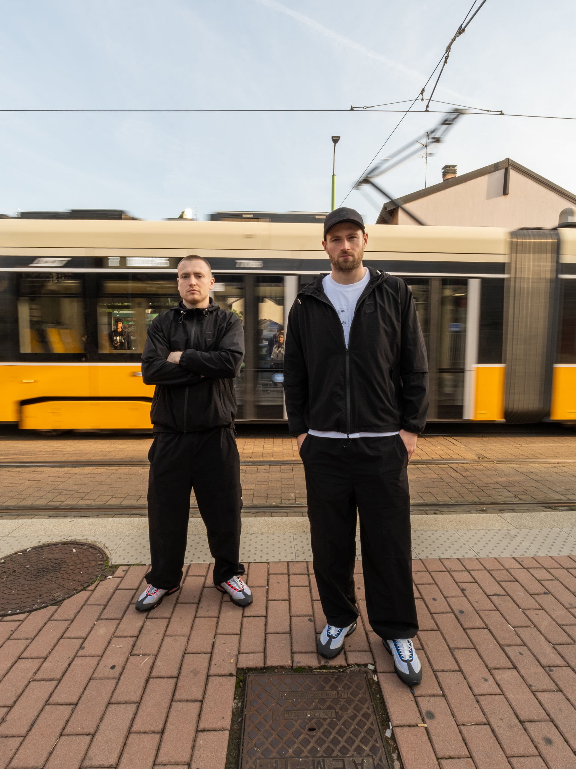 Two men standing on a platform wearing Baggys Vanta Tracksuit, with a tram in the background