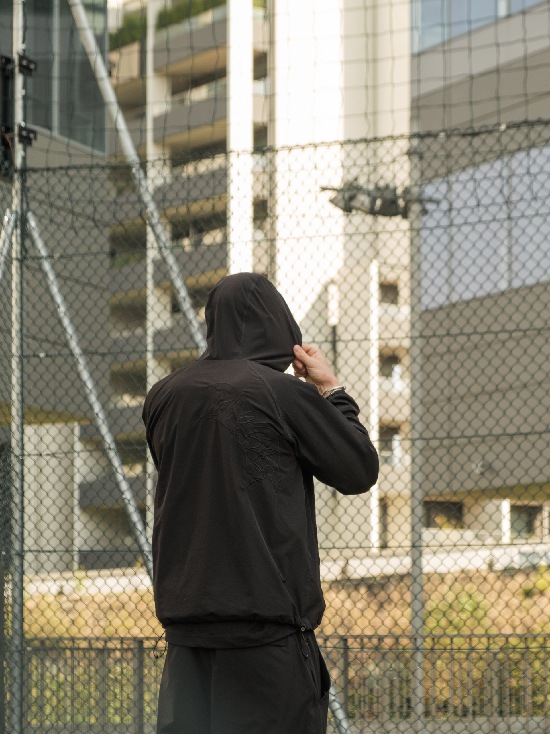 Red wearing Baggys Vanta Tracksuit standing in front of a chain-link fence with buildings in the background