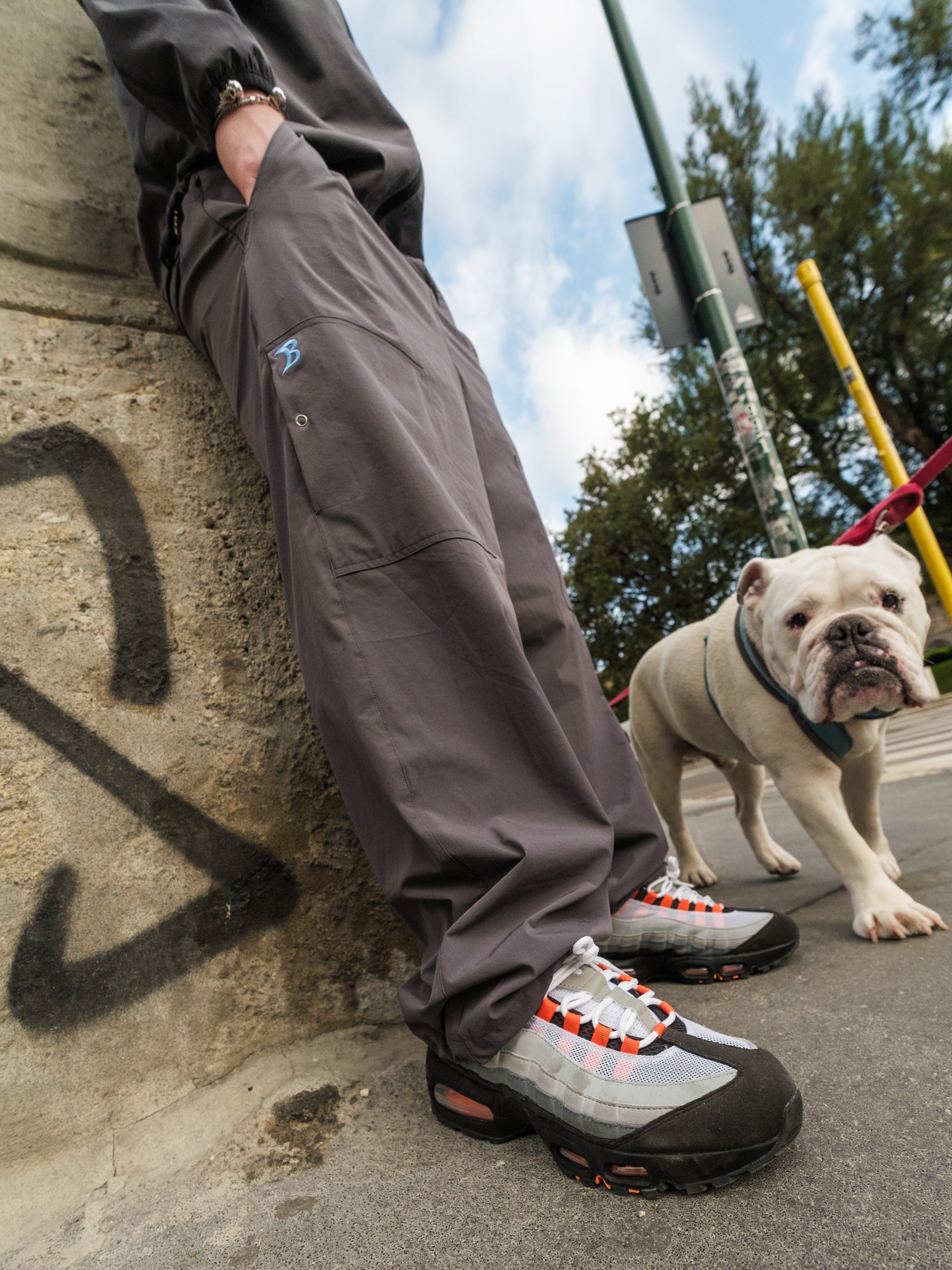 Person wearing Baggys Oxide Track Pants, leaning against a wall with a dog on a leash in the background.