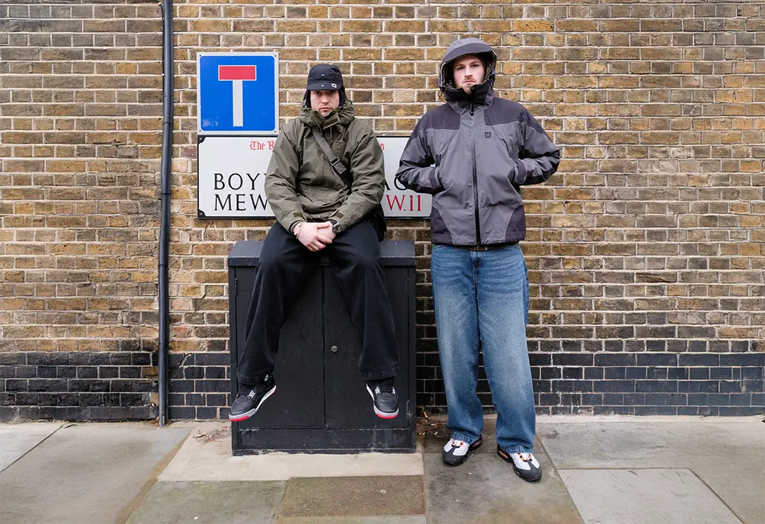 two men against a wall, one with black baggy jeans on, one with blue baggy jeans on