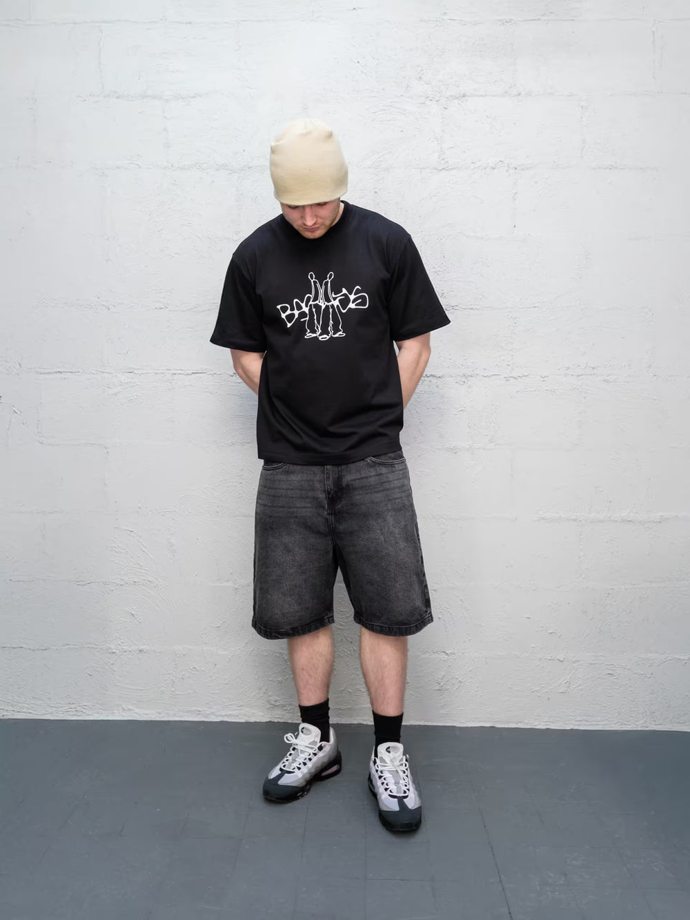 Person wearing a black t-shirt with a graphic design, gray shorts, and a beige beanie against a white brick wall.