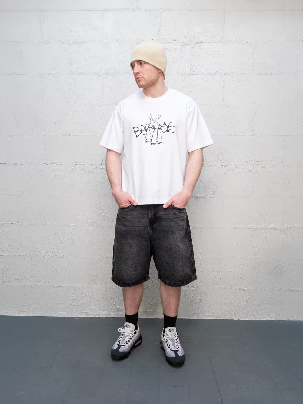 Person wearing a white t-shirt with text, black shorts, and a beige beanie against a gray brick wall.