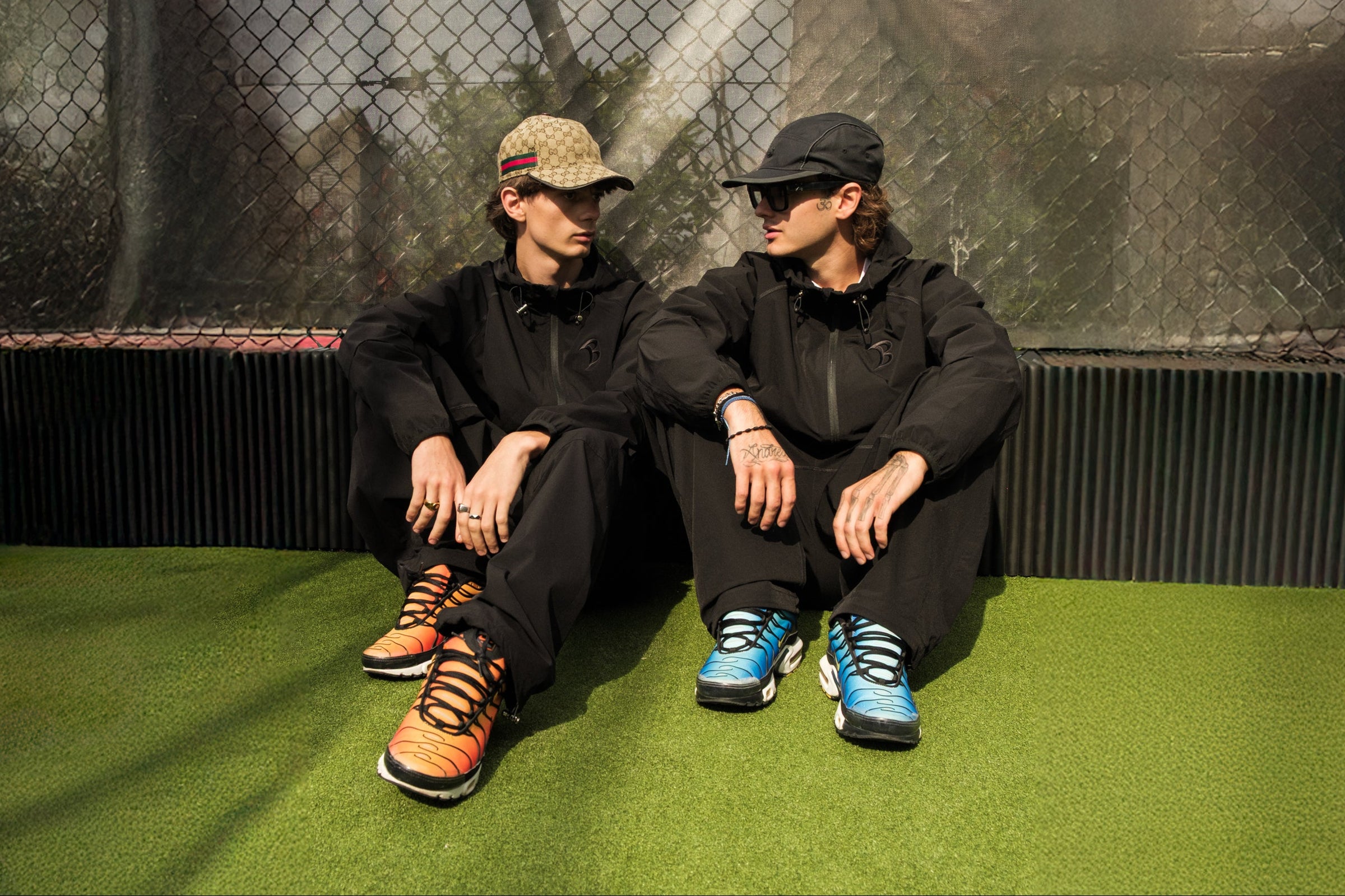 Two people sitting on a green surface wearing black outfits with colorful shoes, in front of a chain-link fence.