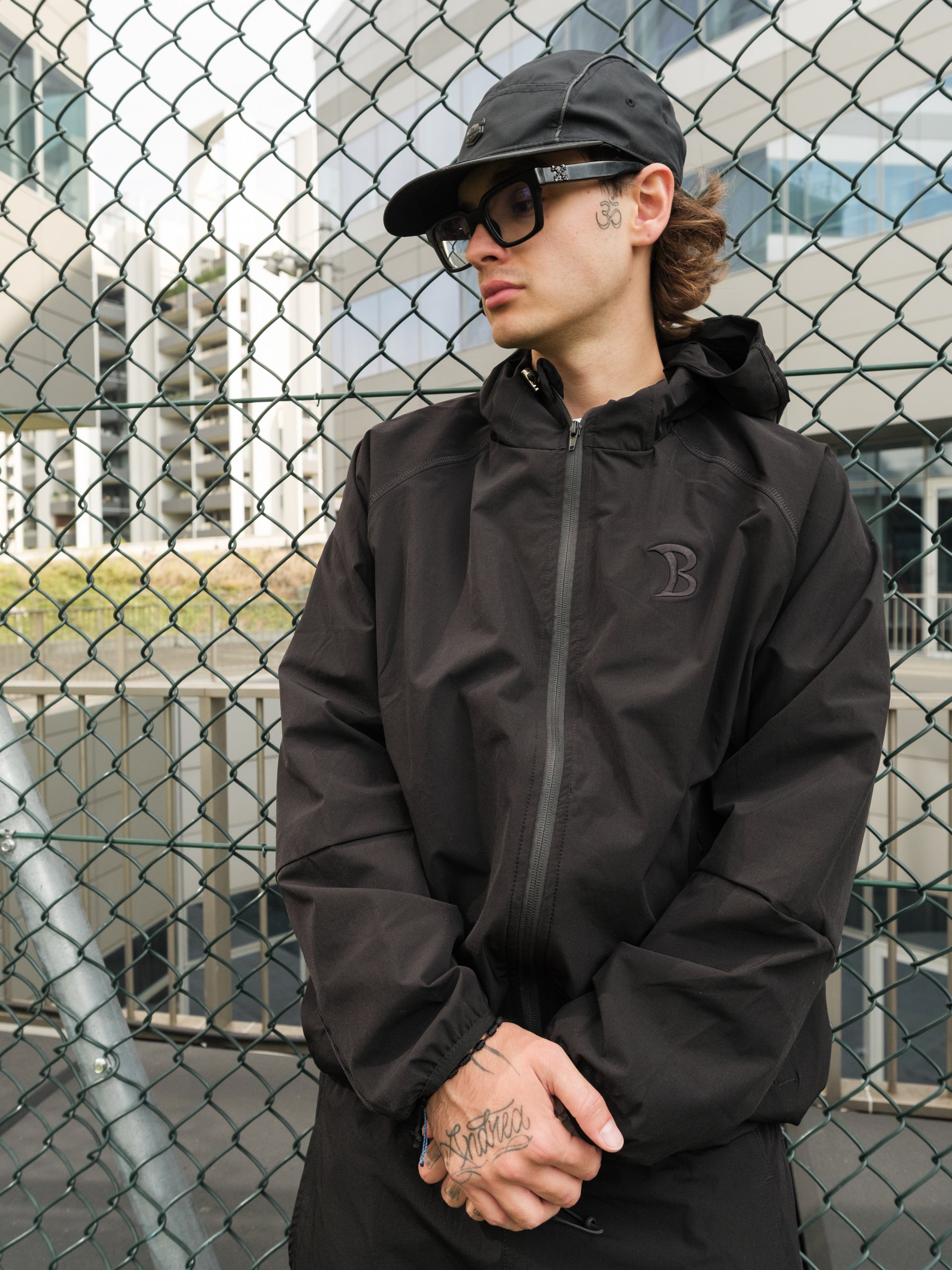 Person wearing a black jacket and cap standing in front of a chain-link fence.