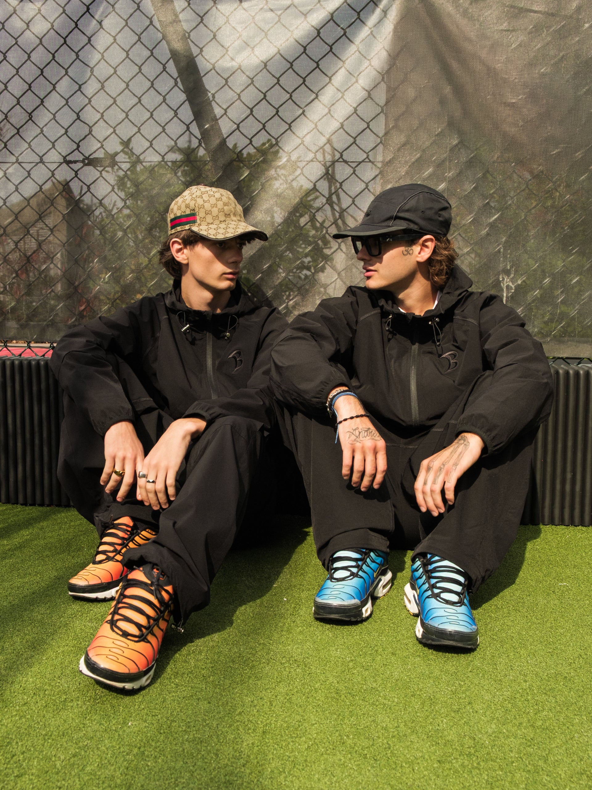 Two people sitting on a grassy area wearing black outfits and colorful sneakers.
