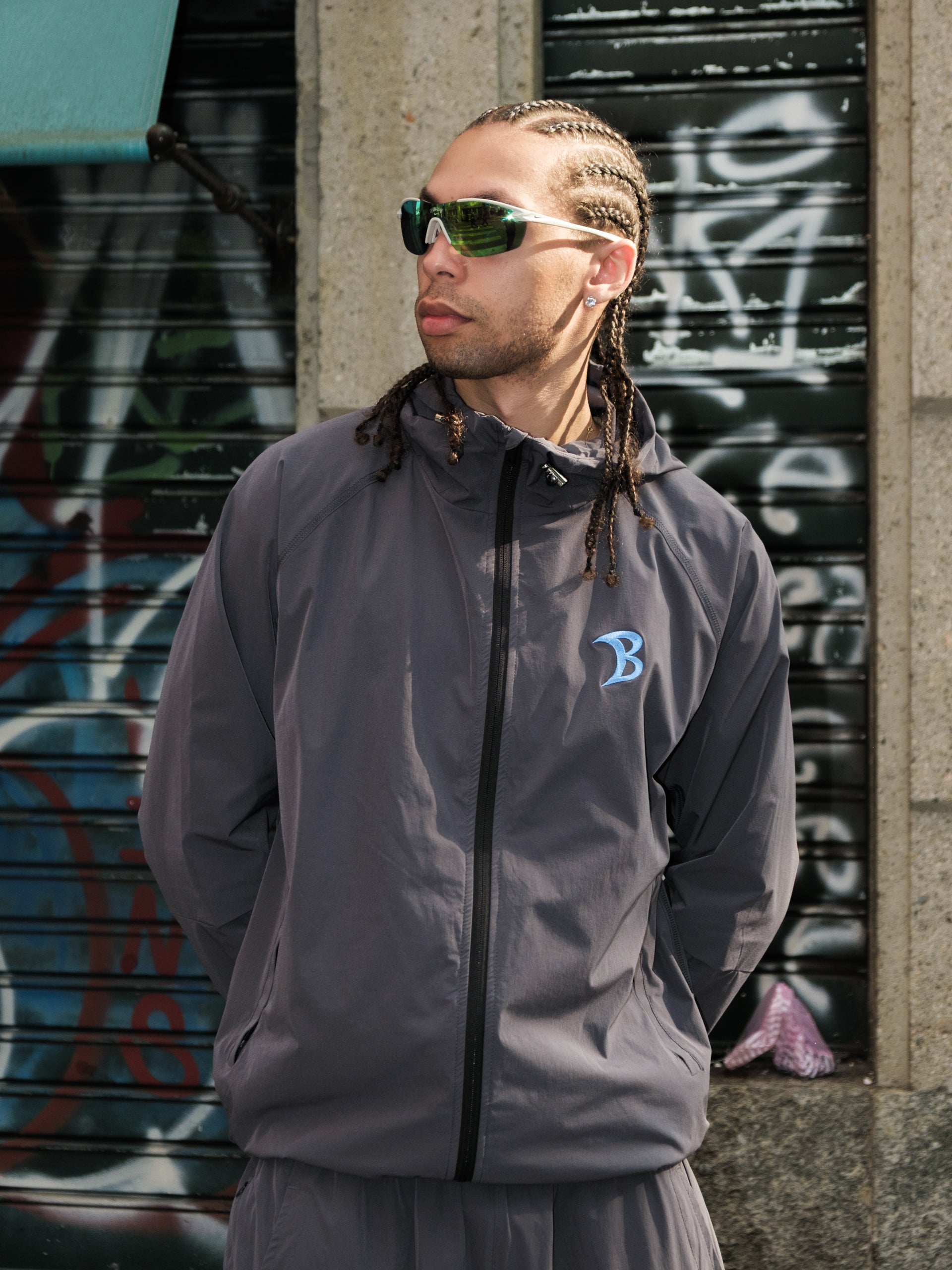 Man wearing a gray jacket with a logo, sunglasses, and braided hair standing against a graffiti-covered wall in Milan