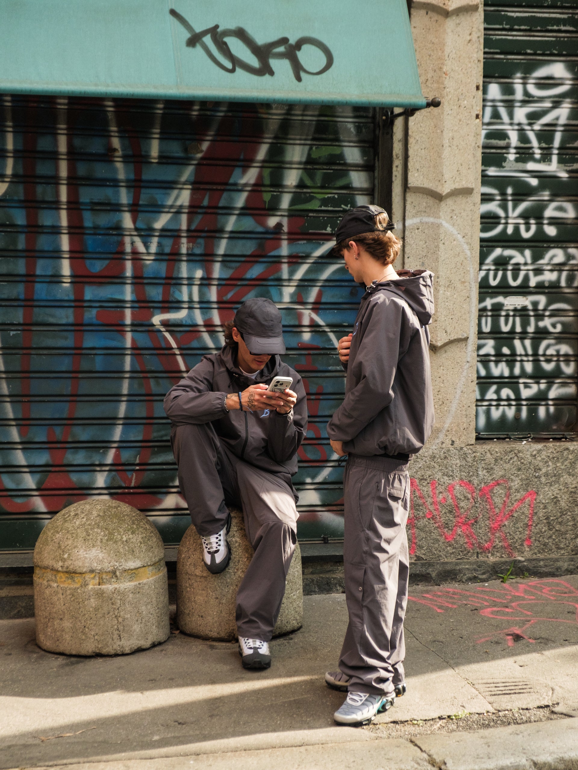 Two people wearing Baggys Oxide Tracksuit using their phones on a street with graffiti-covered walls in Milan 