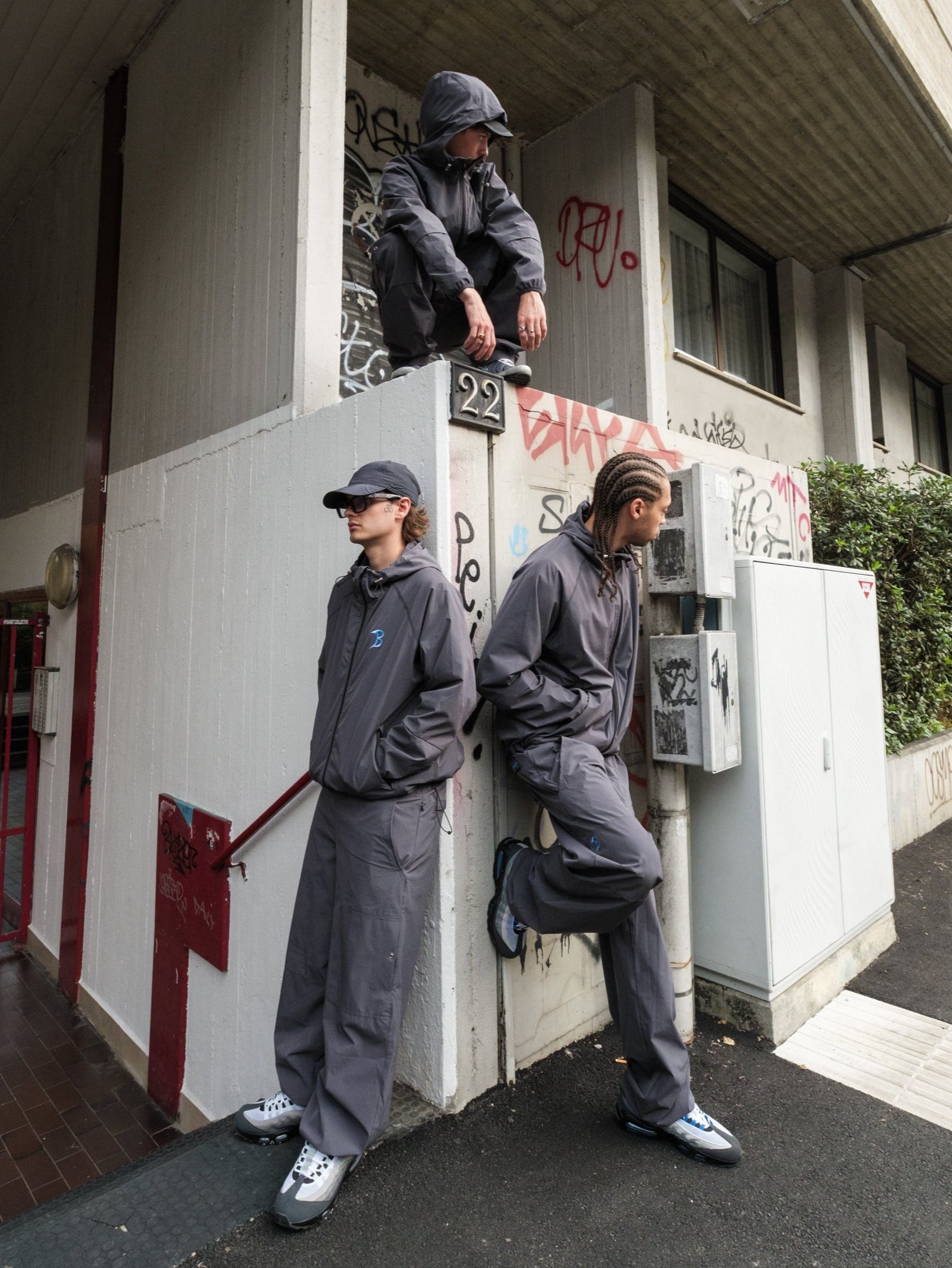 Three people in Baggys Oxide Tracksuit standing on a sidewalk with graffiti on a building.