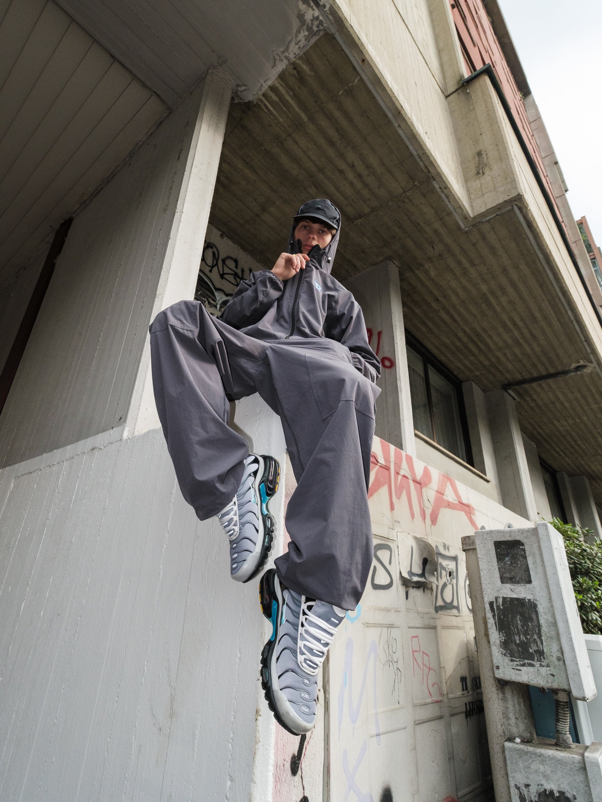 Person in gray tracksuit and sneakers hanging from a building ledge with graffiti.