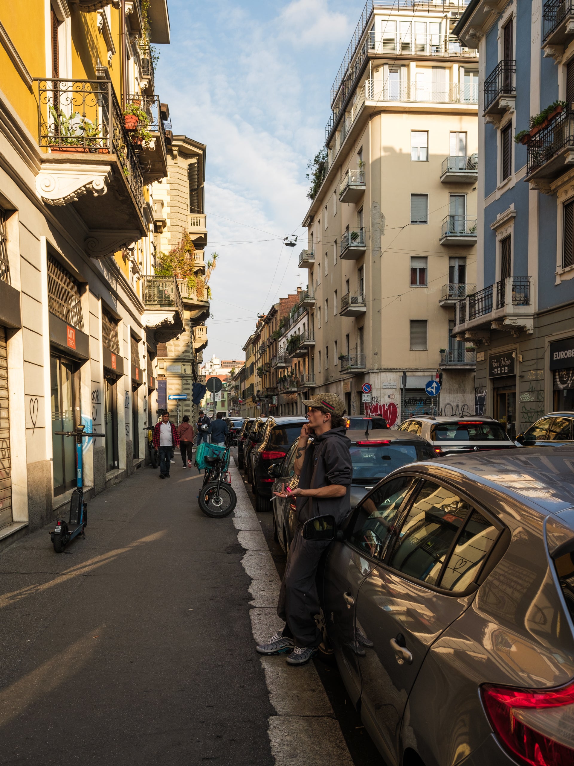 City street scene with buildings, parked cars, and Steffano wearing Baggys Oxide Tracksuit whilst on the phone and having a cigarette 