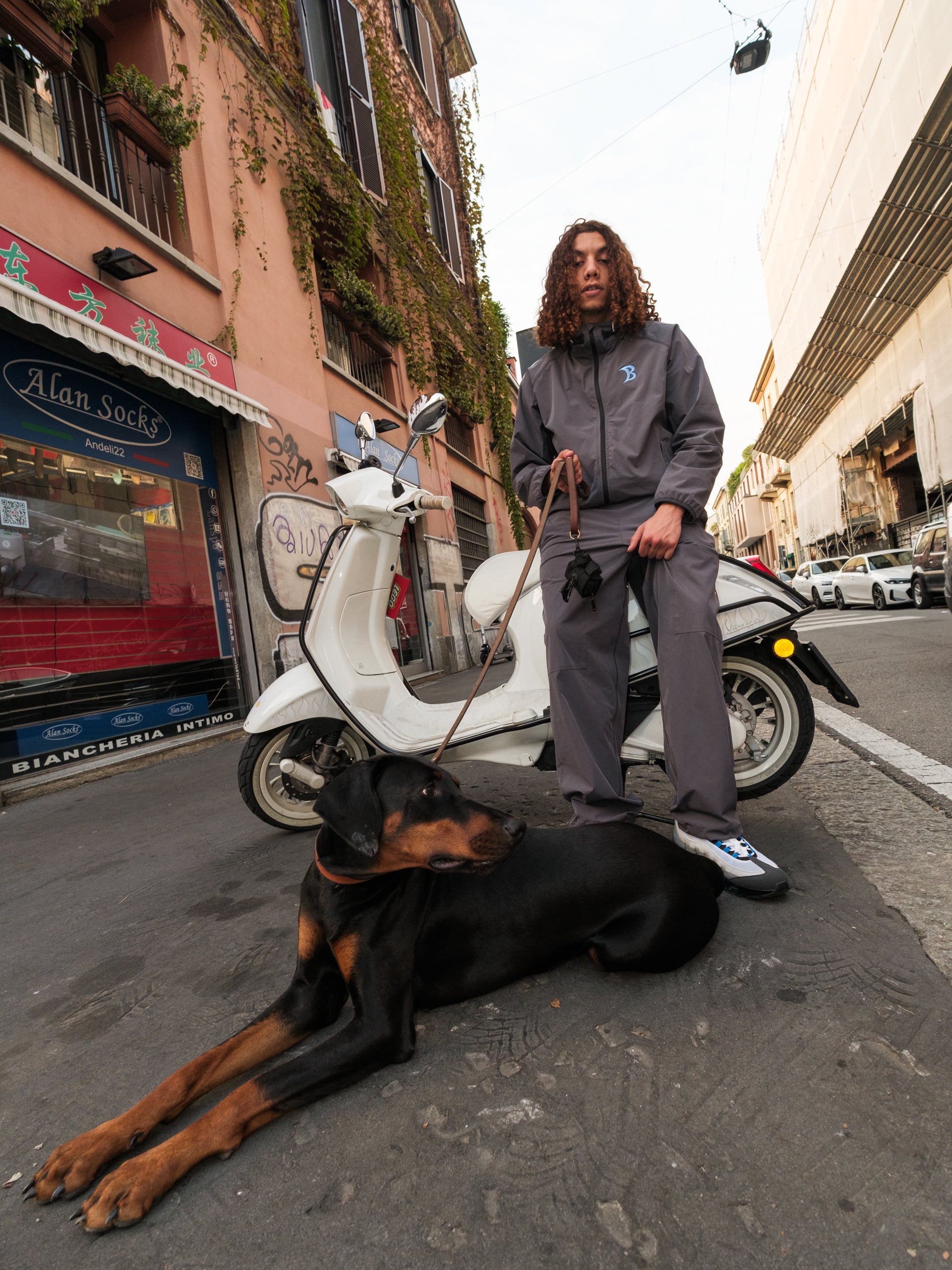 Person standing next to a dog on a leash in an urban street setting with a scooter.