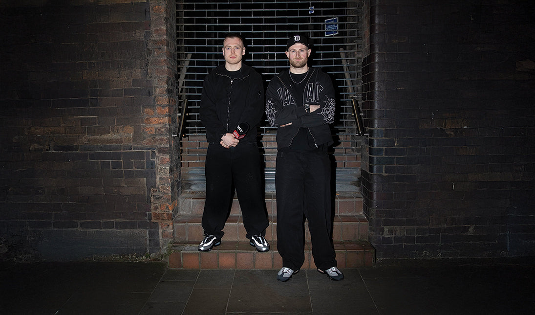 Two men in wearing baggy jeans standing on a staircase against a brick wall.